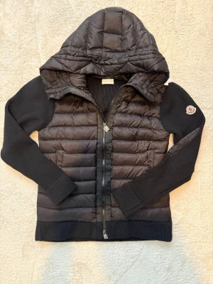 Svart Moncler cardiganjacka med huva - Snygg svart cardiganjacka från Moncler med vadderad framsida och huva. Jackan har dragkedja framtill och ribbade muddar. Kan gå ner i pris vid en snabb affär. Kontakta för mer bilder.
