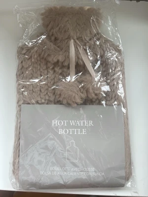 Värmeflaska med beige fluffigt fodral - Säljer en värmeflaska med mjukt, beige fluffigt fodral och dekorativa tofsar. Perfekt för att hålla sig varm under kalla dagar. Fodralet är gjort av syntetmaterial och flaskan är av plast. Levereras i originalförpackning.