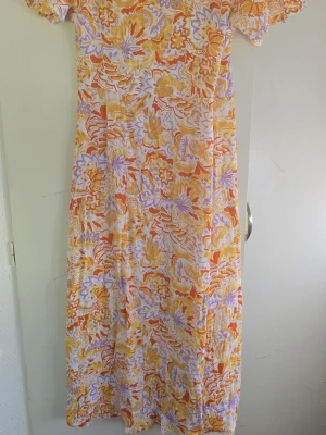 Blommig omlottklänning i pastell - Lång omlottklänning med korta ärmar och blommigt mönster i lila, orange och gul på vit botten. Klänningen har volangkant nertill och knytband i midjan för en feminin siluett. Perfekt för varma dagar och somriga tillfällen.