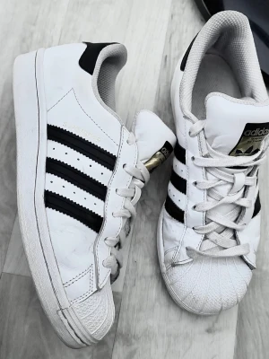 Adidas Superstar vita sneakers - Klassiska Adidas Superstar sneakers i vitt läder med svarta ränder och guldiga detaljer på plösen. Skorna har den ikoniska snäckformade tån och vita skosnören. Perfekta för en streetwear-look och passar till det mesta.