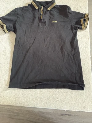 Svart pikétröja från BOSS - Svart pikétröja från BOSS med guldfärgade detaljer på krage och ärmslut. Tröjan har korta ärmar, klassisk krage och knappar framtill. BOSS-logga broderad på bröstet. Perfekt för en stilren och sportig look.