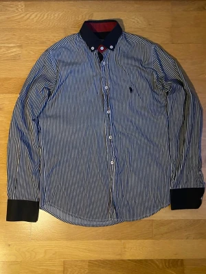 Randig skjorta från Polo Ralph Lauren skickar inom 24h - Säljer en klassisk blå- och vitrandig skjorta från Polo Ralph Lauren med lång ärm och knappar framtill. Skjortan har mörkblå krage och manschetter samt den ikoniska broderade loggan på bröstet. Perfekt för en stilren look. Fattas en knapp. Skickar inom 24h
