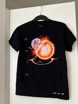 The Weeknd After Hours Tour T-shirt - Säljer en svart t-shirt från The Weeknd med tryck från After Hours Til Dawn Global Stadium Tour. Framsidan har ett coolt rymdmotiv med exploderande planeter och texten AHTD. Baksidan listar turnéstopp. Perfekt för fans av The Weeknd och snygg till streetwear.