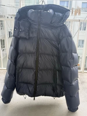 Svart dunjacka från J.Lindeberg - Snygg svart dunjacka från J.Lindeberg med hög krage, avtagbar huva och två dragkedjefickor. Jackan har en glansig finish och breda kanaler fyllda med dun, perfekt för kalla vinterdagar. Innerfodret har diskret logomönster. Storlek M och säljer då ja inte behöver den längre. Köpte för 5500kr och säljer nu billigare. Skicka prisförslag så går de säkert att lösa något;)