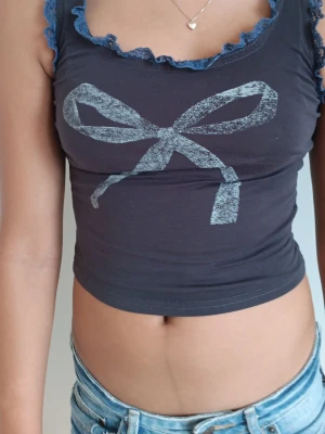 Mörkblå croppad linnetopp med rosett - Snygg mörkblå croppad linnetopp med ljusblå spetsdetaljer vid urringningen och ett stort tryck av en rosett framtill. Perfekt för dig som gillar en feminin och trendig stil. Passar till både jeans och kjol!
