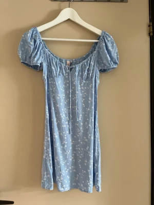 Blommig blå kortklänning från Nelly - Supersöt ljusblå kortklänning från Nelly med puffärmar och små vita blommor. Klänningen har snörning vid urringningen och är gjord i ett lätt material, perfekt för varma dagar. Passar dig som gillar romantisk och trendig stil. Klänningen är använd få gånger men har lite fläckar på baksidan men inget man tänker på💕