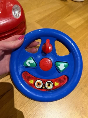Blått leksaksratt till bil - Säljer ett blått leksaksratt i plast med röda och gröna knappar samt dekaler som visar hastighetsmätare. Passar till leksaksbil, perfekt för barn som gillar att låtsasköra. Ratten har en rund form och färgglada detaljer.