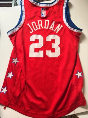 Röd NBA Jordan basketlinne - Säljer ett rött NBA basketlinne med Jordan och nummer 23 på ryggen. Linnet har vita och blå detaljer, stjärnor på sidorna och broderade NBA-loggor. Perfekt för basket eller streetwear. Tillverkad i meshmaterial för bra ventilation.
