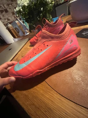 Röda Nike fotbollsskor med blå detaljer - Säljer ett par röda Nike grusfotbollsskor med stor ljusblå swoosh på sidan och blå detaljer vid hälen. Skorna har strumpliknande skaft och är tillverkade i syntetmaterial för bra passform och grepp på planen. Perfekta för dig som vill ha stilrena och funktionella skor till matchen. NIKE AIR ZOOM MERCURIAL VAPOR 16 ACADEMY TF PRISM, storlek 38