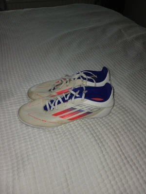 Vita Adidas fotbollsskor med blå detaljer - Säljer ett par vita Adidas fotbollsskor med blå och röda detaljer. Skorna har klassisk snörning, låg profil och är tillverkade i syntetmaterial. Perfekta för dig som vill ha stilrena och lätta skor på planen.