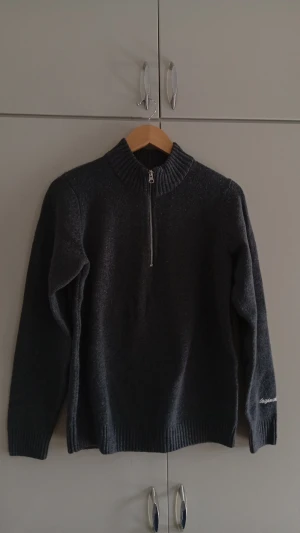 Half zip - Mörkgrå stickad tröja från Jack & Jones halv dragkedja framtill och ribbstickad krage, ärmslut och nederkant. Tröjan har lång ärm och en diskret broderad logga på ena ärmen. Perfekt för kyliga dagar.