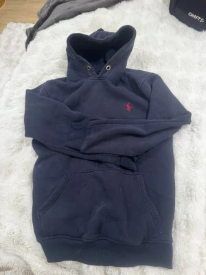 Mörkblå hoodie från Polo Ralph Lauren - Mörkblå hoodie med klassisk röd Polo Ralph Lauren-logga broderad på bröstet. Hoodien har känguruficka, ribbade muddar och metallöljetter vid huvan. Perfekt för en avslappnad och stilren look.