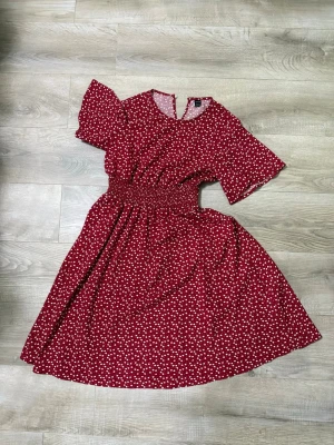 Röd blommig midiklänning - Söt röd klänning med vita små blommor, kort ärm och smock i midjan som ger en fin siluett. Kjolen har en lätt utställd form och tyget känns luftigt, perfekt för varma dagar. Rund halsringning och bekväm passform.