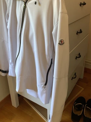 Vit vindjacka från Moncler - Snygg vit vindjacka från Moncler med huva och hel dragkedja framtill. Jackan har svarta detaljer vid fickor och ärmslut samt Moncler-logga på ärmen. Tillverkad i ett lätt och smidigt material som passar perfekt för blåsiga dagar.