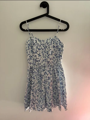 Vit och blå blommig sommarklänning - Söt kort klänning med tunna axelband och blått blommönster på vit botten. Klänningen har en lätt v-ringad urringning och smock i ryggen för skön passform. Perfekt för varma dagar och enkel att styla med sneakers eller sandaler.