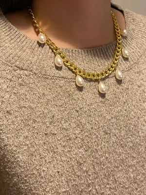 Guldhalsband med droppformade pärlor - Snyggt halsband i guldfärgad med droppformade vita pärlor som hänger längs hela kedjan. Kedjan är chunky och ger ett trendigt intryck, perfekt för att lyfta vilken outfit som helst. Materialet är rostfritt.