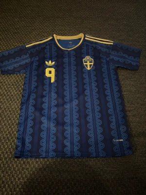 Blå Sverige fotbollströja Adidas Isak M - Säljer en blå fotbollströja från Adidas med Sveriges landslagsemblem och nummer 9 i gult. Tröjan har retroinspirerat mönster, gula detaljer på krage och axlar samt klassiska Adidas-ränder. Perfekt för match eller träning.