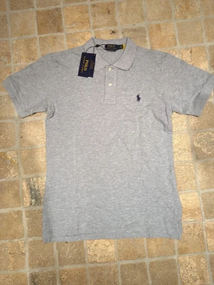 Grå pikétröja från Polo Ralph Lauren - Snygg grå pikétröja från Polo Ralph Lauren med klassisk krage och två knappar framtill. Tröjan har korta ärmar och den ikoniska broderade loggan på bröstet. Perfekt till jeans eller shorts för en clean och stilren look.