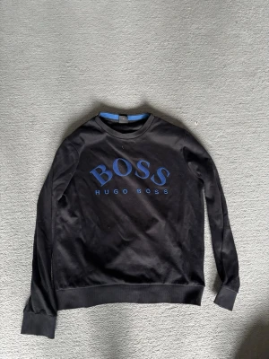 Svart sweatshirt från Hugo Boss - Snygg svart sweatshirt från Hugo Boss med stort blått BOSS-tryck på bröstet. Tröjan har rund halsringning, ribbade muddar vid ärmslut och nederkant samt en avslappnad passform. Perfekt för en stilren och casual look.