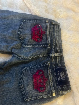 Victoria Beckham  jeans med rosa  - Säljer ett par blå jeans från Victoria Beckham med raka ben och snygga rosa kronbroderier på bakfickorna. Jeansen har klassisk femficksmodell, gylf med dragkedja och knapp samt detaljer. Materialet är denim i bomull. De står W30 i dem så ungefär storlek EU 38.