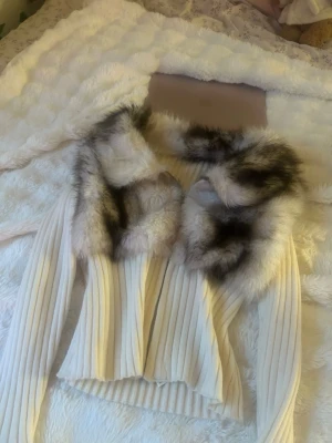 Vit ribbad pälskofta med fuskpäls - Supersnygg vit ribbad kofta med bred krage i fluffig fuskpäls i grå och bruna toner. Västen har en figurnära passform och ger en lyxig känsla till din outfit. Perfekt för dig som vill sticka ut med en trendig look och älskar detaljer i fuskpäls.