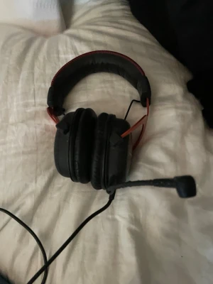 HyperX gaming headset med mikrofon - Säljer ett HyperX gaming headset med sladd och inbyggd mikrofon. Hörlurarna är svarta med röda detaljer och har mjuka, vadderade kåpor samt justerbart huvudband. Perfekt för gaming och voice chat. Klassisk over-ear design med robust metallbåge.