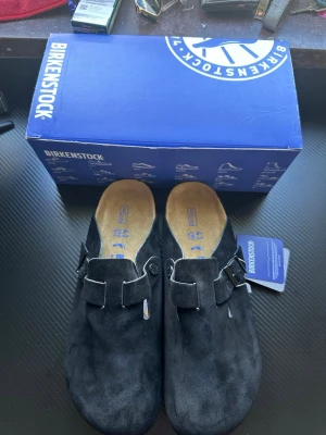 Svarta Birkenstock tofflor i mocka - Säljer ett par svarta Birkenstock tofflor i mjuk mocka med justerbart spänne och korksula. Klassisk slip-in modell med bekväm innersula och diskret Birkenstock-logga på sidan. Perfekta för dig som gillar stilrena och sköna skor.