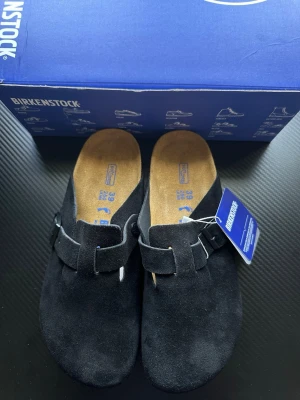 Svarta Birkenstock tofflor i mocka - Säljer ett par svarta Birkenstock tofflor i mjuk mocka med justerbart spänne och korksula. Klassisk slip-in modell med bekväm fotbädd och diskret logga på sidan. Perfekta för dig som gillar stilrena och tidlösa skor.
