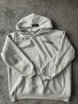Grå hoodie från Essentials Fear of God - Snygg grå hoodie från Essentials Fear of God med svart logga på bröstet och patch på huvan. Klassisk passform med känguruficka och ribbade muddar. Tillverkad i mjukt bomullsmaterial som känns riktigt skönt mot huden.
