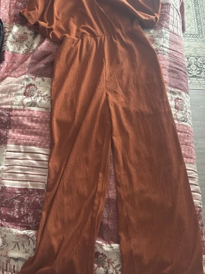 Rostbrun vid jumpsuit med resårmidja - Säljer en snygg rostbrun jumpsuit med vida ben och resår i midjan. Materialet är mjukt och har en lätt struktur som ger en avslappnad känsla. Perfekt för dig som gillar en enkel men trendig stil. Inga synliga fickor eller detaljer.