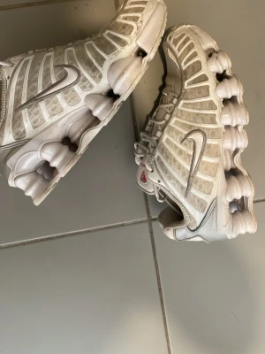 Vita Nike Shox sneakers - Nike Shox sneakers i vitt med silverdetaljer och ikonisk fjädrande sula. Skorna har ett sportigt och futuristiskt utseende med mesh och syntetmaterial i ovandelen samt tydliga Nike-loggor på sidorna. Äkta 