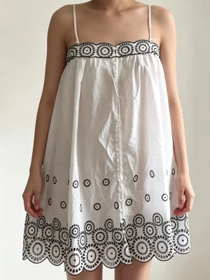 Vit kort klänning med broderi - Söt vit kort klänning med tunna axelband och svart cirkulärt broderi längs överkant och nederkant. Klänningen har en luftig passform och är perfekt för varma dagar. Materialet känns lätt och somrigt.