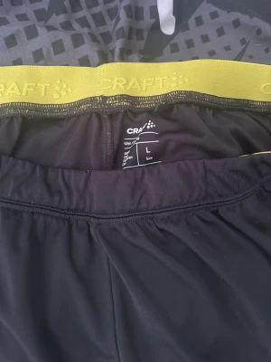 Svarta träningsshorts från Craft L - Säljer ett par svarta träningsshorts från Craft med gul resår i midjan och IFK-logga på ena benet. Shortsen är tillverkade i ett lätt och snabbtorkande material, perfekta för sport och aktivitet. Diskret Craft-logga på benet.