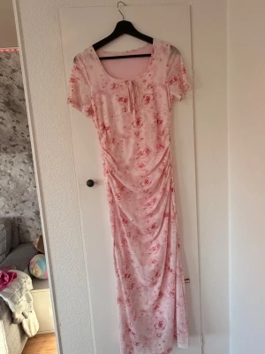 Ljusrosa långklänning med rosor -  Rosa klänning med blommor och t-shirt armar, rosett fram. Märke och storlek oklart. Men gissar på storlek M och märke SHEIN