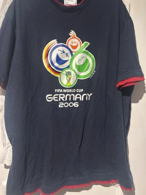 FIFA World Cup Germany 2006 t-shirt - Mörkblå t-shirt med färgglatt FIFA World Cup Germany 2006-tryck på bröstet. T-shirten har röda detaljer vid ärmslut och nederkant samt klassisk rund hals. Perfekt för dig som gillar fotboll och retrostil.