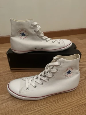 Vita Converse All Star high tops helt nya - Klassiska vita Converse All Star high tops med canvasmaterial och vit gummisula. Skorna har den ikoniska loggan på sidan, vita skosnören och röda detaljer längs sulan. Perfekta för en avslappnad och stilren look. Helt nya. Ordinariepris: 700 kr.