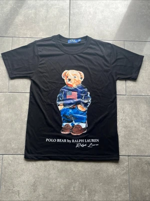 Polo Ralph Lauren Polo Bear T-shirt – Svart | Strl S - Säljer en svart Polo Ralph Lauren T-shirt med det ikoniska Polo Bear-motivet på framsidan. En klassisk och stilren T-shirt i mycket fint skick.  * Storlek: S * Färg: Svart * Skick: Mycket fint skick * Klassiskt Polo Bear-print * Skickas endast. Hör gärna av dig om du har några frågor!