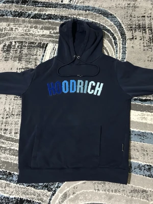 Mörkblå HOODRICH hoodie - Snygg mörkblå hoodie från HOODRICH med stor broderad logga i blå och vita nyanser på bröstet. Klassisk känguruficka framtill och justerbar huva med dragsko. Perfekt för en chill och street-inspirerad look.