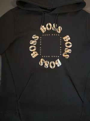 Svart hoodie från Hugo Boss - Snygg svart hoodie från Hugo Boss med stor grafisk BOSS-logga i beige och vitt på bröstet. Klassisk huva med dragsko och känguruficka framtill. Tillverkad i mjukt material som känns skönt mot huden. Perfekt för en avslappnad och trendig look.