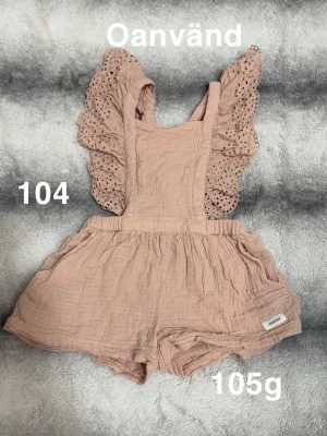 Rosa jumpsuit med volangärmar - Supersöt rosa jumpsuit med breda axelband och detaljerade volangärmar i broderad spets. Resår i midjan för skön passform och korta ben. Perfekt för en trendig och bekväm look.🌸Oanvänd. Storlek 104.