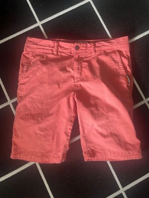 Korallrosa shorts från Superdry - Snygga korallrosa shorts från Superdry med klassisk passform. Shortsen har bälteshällor, knappgylf och fickor både fram och bak. Perfekta för varma dagar och ger en fräsch look till din outfit.