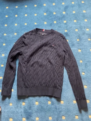 Mörkblå kabelstickad tröja Tommy Hilfiger - Snygg mörkblå kabelstickad tröja från Tommy Hilfiger med rund halsringning och diskret logga på bröstet. Tröjan har långa ärmar och ribbade muddar vid ärmslut och nederkant. Perfekt för lager på lager och klassisk stil.