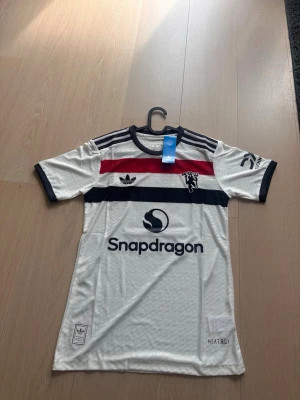 Manchester United bortatröja Adidas - Säljer en vit Manchester United bortatröja från Adidas med svarta och röda detaljer, samt sponsorlogga Snapdragon på bröstet. Tröjan har B. Fernandes och nummer 8 på ryggen. Tillverkad i lätt och ventilerande material med HEAT.RDY-teknologi.