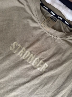 Beige linne topp från Stronger - Snygg beige långärmad topp från Stronger med rund halsringning och tryckt logga framtill. Tillverkad i mjuk bomull som känns skön mot huden. Perfekt för dig som gillar enkel och clean stil.