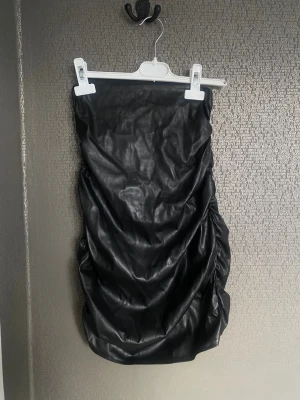 Svart axelbandslös kort klänning - Säljer en svart axelbandslös kort klänning i skinnimitation. Klänningen har en tight passform med rynkade sidor som ger en cool och edgy look. Perfekt för dig som vill sticka ut och gillar trendiga material.