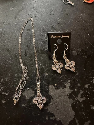 Halsband och örhängen med liljemotiv - Snyggt set med halsband och matchande örhängen i silverfärgad metall. Båda har detaljerade liljemotiv som ger en cool och elegant vibe. Perfekt för dig som gillar unika accessoarer med lite extra edge.