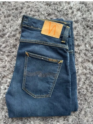 Nudie Jeans grim Tim - Säljer ett par mörkblå jeans från Nudie Jeans med klassisk femficksmodell och kontrastsömmar. Jeansen har en snygg läderpatch med logga bak och vågig detalj på bakfickan. Tillverkade i slitstark denim, perfekta till vardagslooken.