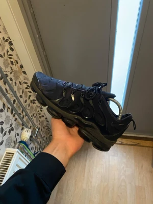 Svarta Nike Air VaporMax Plus - Säljer ett par svarta Nike Air VaporMax Plus sneakers med chunky sula och coola detaljer i plast längs sidorna. Skorna har en sportig vibe med textil och syntetmaterial, samt klassiska snörningar. Perfekta för dig som gillar streetwear och vill sticka ut. Har hål i bubblorna