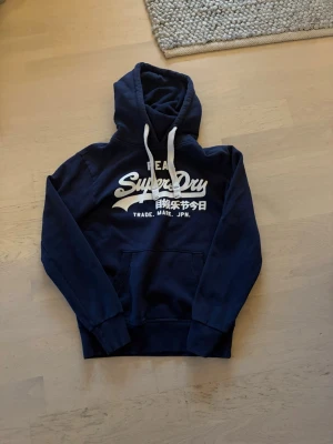 Mörkblå Superdry hoodie med tryck (beskrivning) - Snygg mörkblå hoodie från Superdry med vitt tryck och japanska tecken på bröstet. Klassisk känguruficka framtill och vita dragsnören i huvan. Perfekt för en avslappnad streetstyle-look. säljer gärna med instabox!!