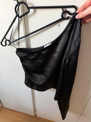 Svart oneshoulder topp från NA-KD - Snygg svart oneshoulder topp från NA-KD i ett glansigt material. Toppen har en lång ärm och sitter tight över kroppen, perfekt för en trendig look. Enkel att matcha med både jeans och kjol.